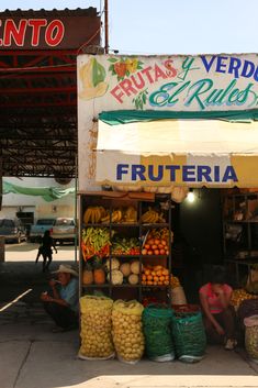 a fruit and vegetable stand in front of a building with people sitting under the awning