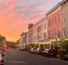 cars parked on the street in front of some buildings at sunset or dawn with pink clouds