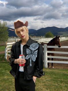 a person standing in front of a fence with a horse behind them and a drink in hand