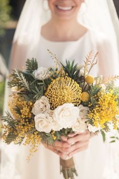 a bride holding a bouquet of yellow and white flowers in her hands, smiling at the camera