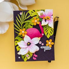 a pink flamingo with tropical leaves and flowers on a purple card next to a white flower