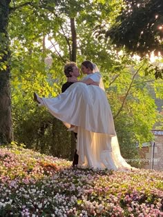 a bride carrying her groom in the middle of a flower garden with trees and flowers all around