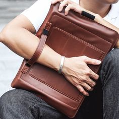 a man sitting on the ground holding a brown leather bag with his hands folded over it