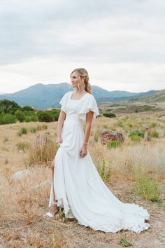 a woman wearing a white dress standing in the middle of a field with mountains behind her