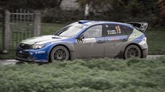 a subaru racing car driving on a road near some bushes and grass in front of a fence