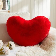 a red heart shaped pillow sitting on top of a white couch next to some flowers