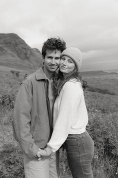 a man and woman standing next to each other in the grass with mountains behind them