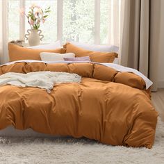 an orange comforter on a bed in front of a window with curtains and flowers