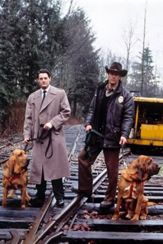 two men standing on train tracks with dogs in front of them and a yellow truck behind them