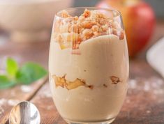 a dessert in a glass on top of a wooden table next to a spoon and an apple