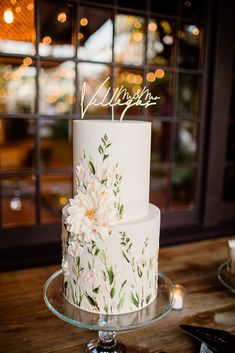 a white wedding cake with flowers and the word happily written on top is sitting on a table in front of a window