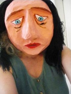 a woman with long black hair and blue eyes is wearing a paper mache mask