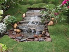 a small waterfall in the middle of a garden filled with rocks and grass, surrounded by pink flowers