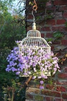 a white birdcage with purple flowers hanging from it's side next to a brick wall