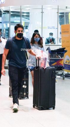 a man and woman walking through an airport with luggage wearing masks on their faces as they walk towards the camera