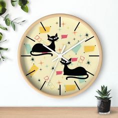 a clock with two cats on it next to a potted plant and a white wall