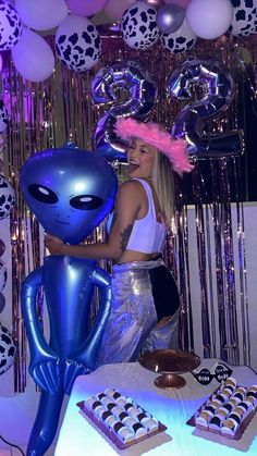 a woman holding an alien balloon next to a table with desserts and balloons on it