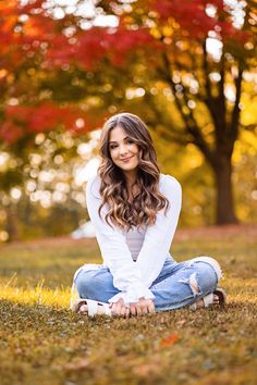 a beautiful young woman sitting on the ground in front of trees with fall foliages