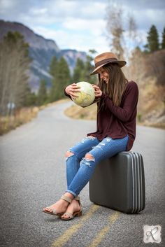 a woman sitting on top of a suitcase while holding a ball and wearing a hat