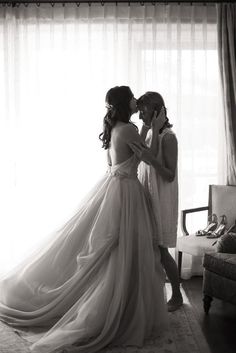 two women in wedding dresses standing next to each other near a window with sheer curtains