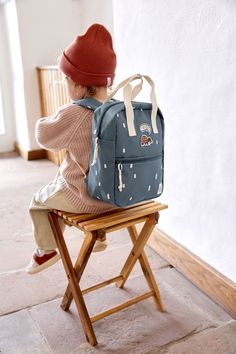 a little boy sitting on a chair with a blue backpack in front of him and wearing a red hat