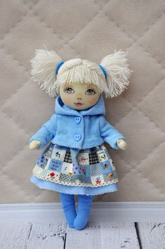 a doll with blonde hair wearing a blue coat and dress is standing against a quilted wall