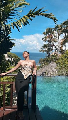 a woman sitting on the edge of a wooden deck next to a swimming pool and palm trees