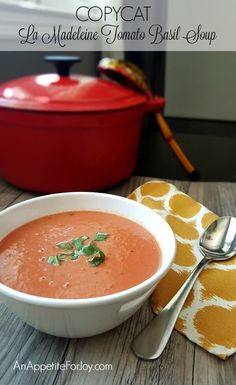 a bowl of tomato soup on a wooden table with a red casserole dish in the background