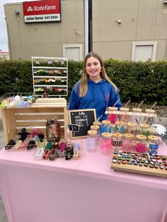 a woman standing behind a pink table with bottles and jars on it in front of a store