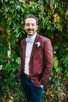 a man in a red jacket and bow tie standing next to some green trees with leaves