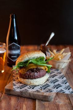 a hamburger sitting on top of a wooden cutting board next to a glass of beer