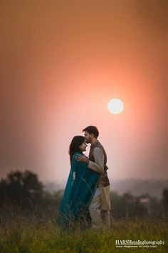 a man and woman standing next to each other in a field with the sun setting behind them