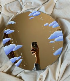 a hand holding up a camera in front of a round mirror with clouds on it