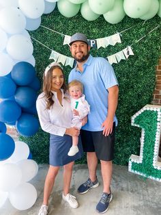 a man and woman holding a baby standing in front of a backdrop with blue, green and white balloons