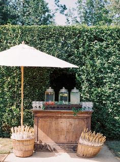 an umbrella and some drinks on a table in front of a bush with bushes behind it