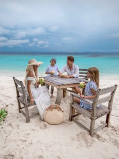 a group of people sitting around a wooden table on top of a sandy beach next to the ocean