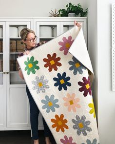 a woman is holding up a quilt in front of a bookcase with flowers on it