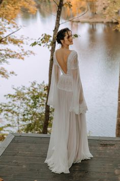 a woman in a wedding dress standing on a deck next to trees looking at the water