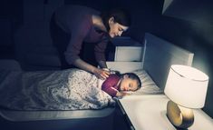 a woman standing over a child laying on a bed next to a night stand and lamp