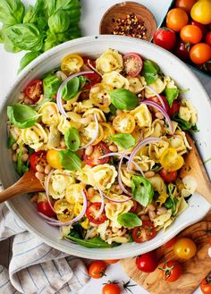 a white bowl filled with pasta salad next to tomatoes, onions and spinach leaves