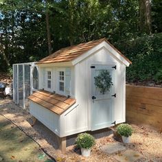 a small white chicken coop in the middle of a yard with trees and bushes around it