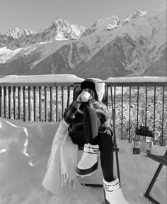 a woman sitting on top of a chair in the snow drinking from a cup with mountains in the background