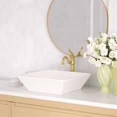 a white sink sitting under a mirror next to a vase with flowers in it and a gold faucet