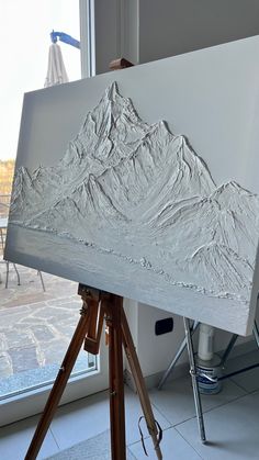 a painting is being displayed on a tripod in front of a window with a view of the mountains