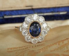 I am thrilled to have acquired this sensational vintage cluster ring for my store. Victorian in style, this piece is graded 'Twentieth Century' by the Guild of Valuers & Jewellers... and I would date this ring as circa 1960. The principal stone is an oval cut natural sapphire surrounded by 10 round brilliant cut diamonds. The sapphire is an impressive 1.06ct and the colour of the stone is graded 'purplish blue', which is a very desirable shade.  The bright, cloudy light was ideal to capture a tr Vintage Blue Sapphire Ring With Halo Setting, Vintage Sapphire Ring With Halo Setting, Vintage Sapphire Diamond Ring With Halo Setting, Vintage Sapphire Cluster Ring With Gemstones, Vintage Blue Brilliant Cut Cluster Ring, Classic Sapphire Diamond Cluster Ring, Vintage Blue Cluster Ring With Brilliant Cut, Vintage Sapphire Diamond Formal Ring, Vintage Sapphire Diamond Ring For Formal Events