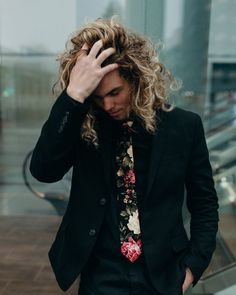 a man with curly hair wearing a black suit and floral neck tie standing in front of an escalator