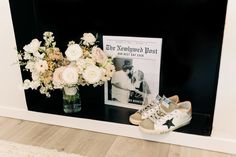 a vase with white flowers and baby's shoes next to a magazine on a shelf