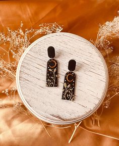 two black and gold earrings sitting on top of a white circular table next to dried flowers