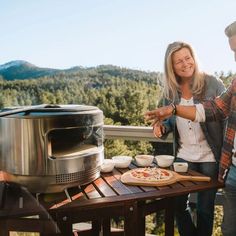 a woman standing next to a pizza on top of a wooden table near a grill