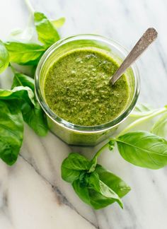 a glass cup filled with green pesto next to lettuce and basil leaves